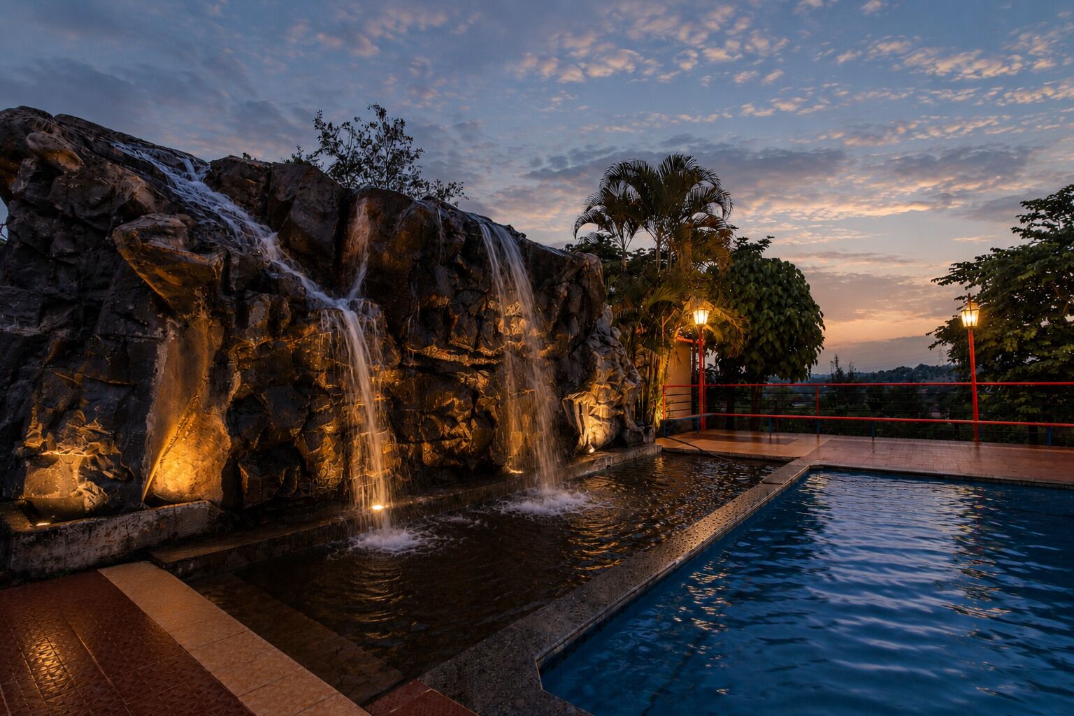 A pool with a waterfall feature, surrounded by rocks and palm trees, under a twilight sky.