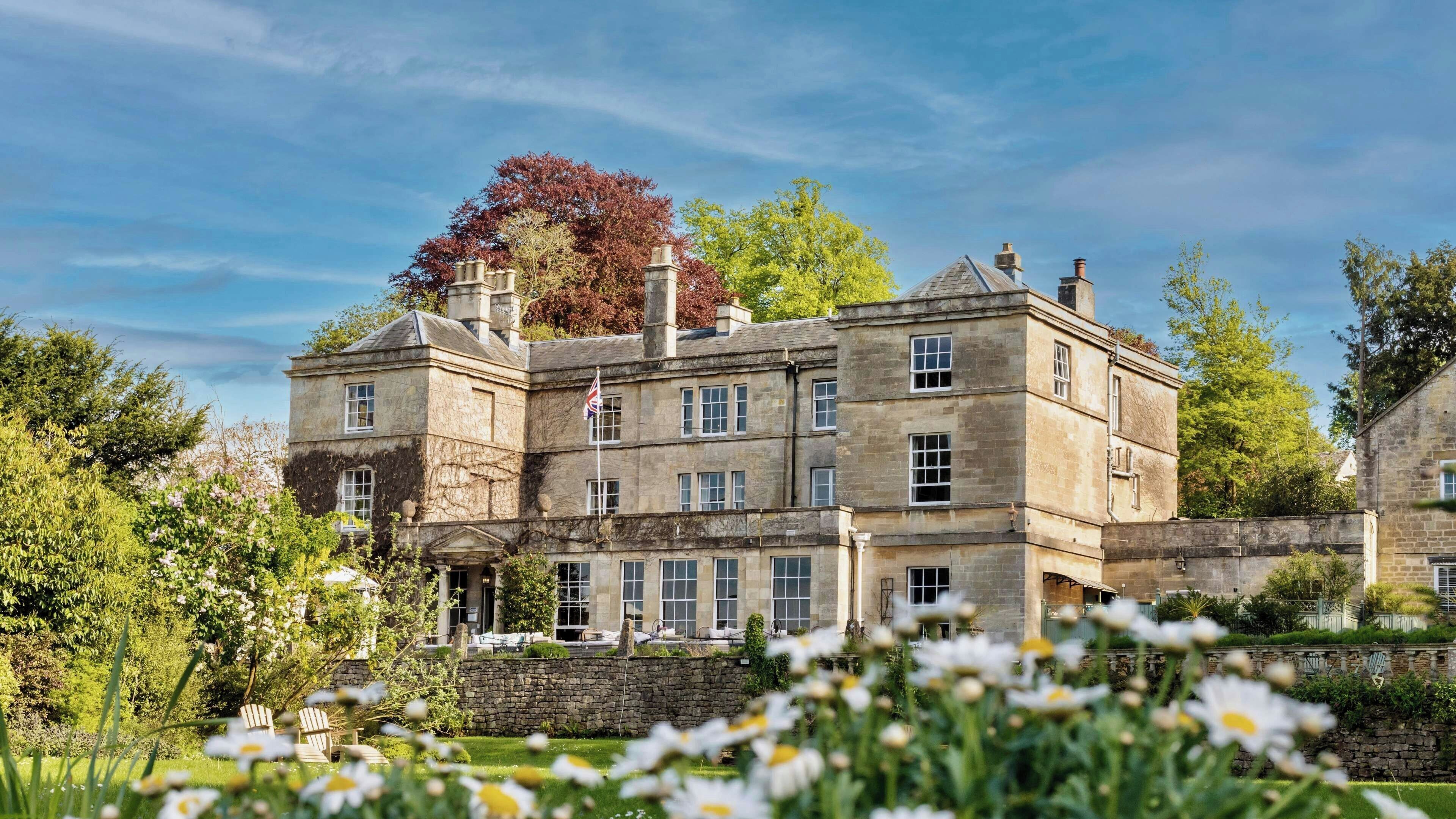 A large stone building with multiple windows and a flag, surrounded by greenery and flowers.