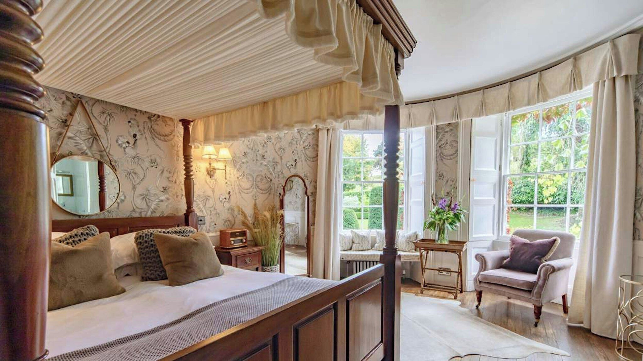 A bedroom with a four-poster bed, a sitting area with a chair and table, and a view of the garden through large windows.