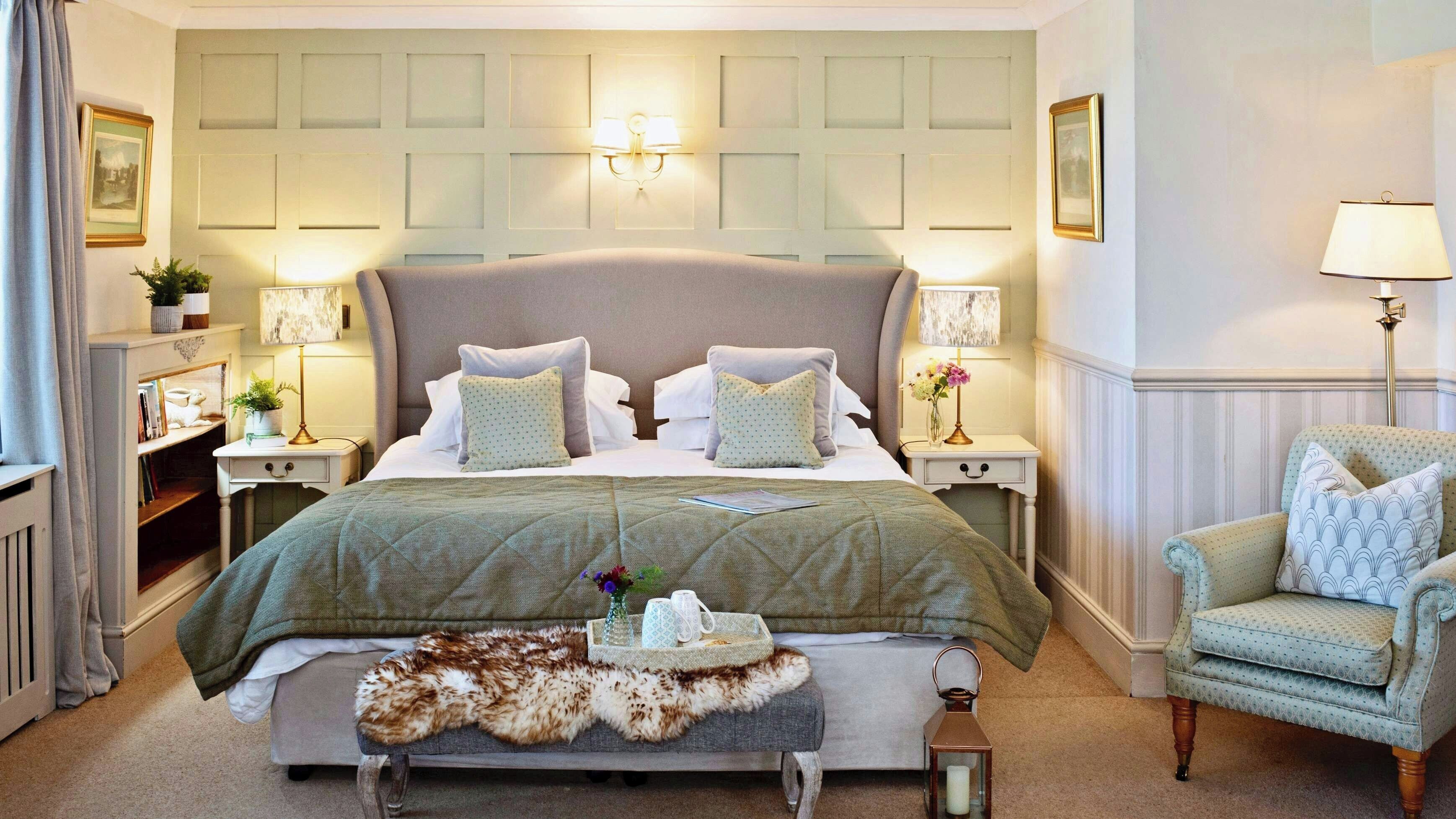 Une chambre élégamment aménagée, dotée d’un grand lit, de tables de chevet avec des lampes, d’un fauteuil et d’un plaid décoratif en fourrure.
