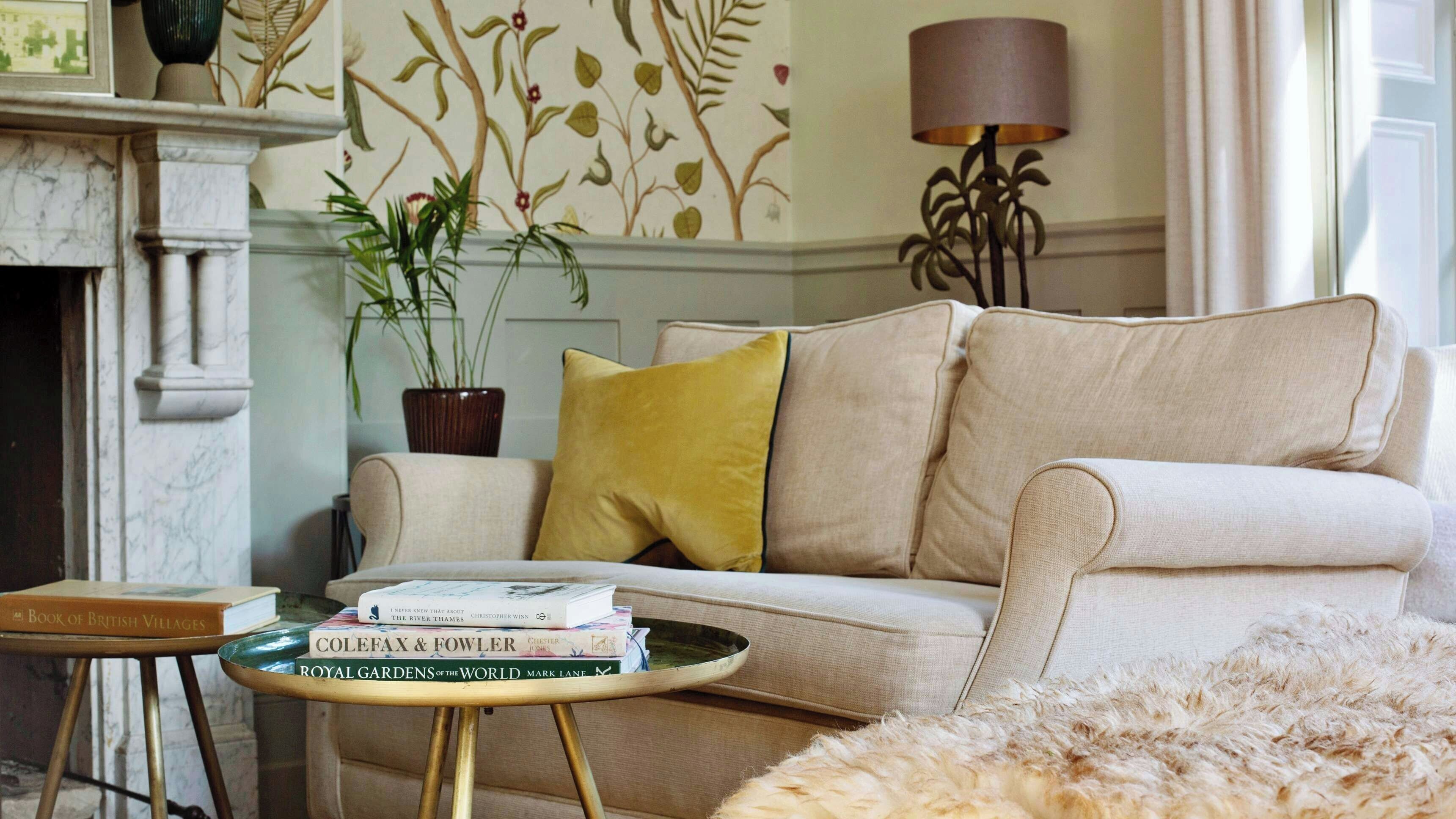 A living room with a beige sofa, a small round table with books, a potted plant, and a lamp.