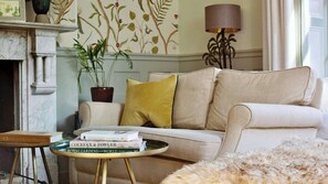Una sala de estar con un sofá color beige, una mesita redonda con libros, una planta en maceta y una lámpara.