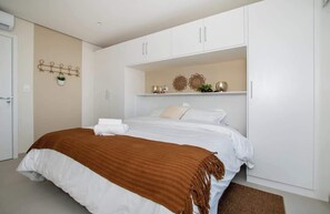 Un dormitorio con cama, armarios blancos, una planta colgada en la pared y objetos decorativos.