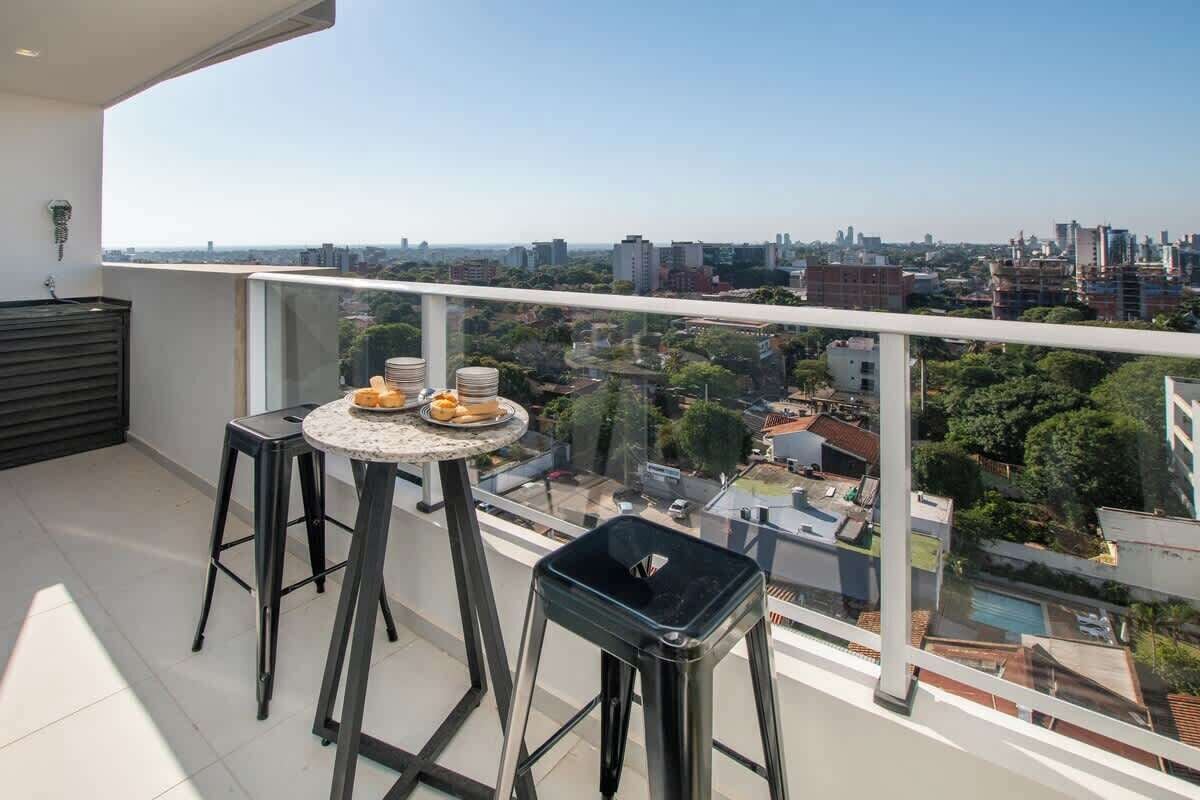 Un balcón con una mesa, dos taburetes y vistas a un paisaje urbano.