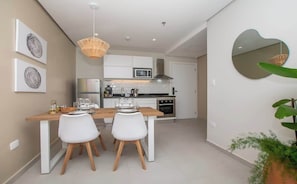 Una cocina moderna con armarios blancos, microondas y estufa. Hay una zona de comedor con una mesa de madera y sillas blancas.