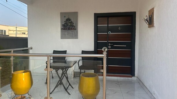 A balcony with a glass railing, a dining table with chairs, and two yellow vases. - BGG Havens (Accra)