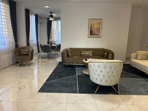 A modern living room with a brown sofa, a white armchair, and a dining area in the background. - BGG Havens (Accra)