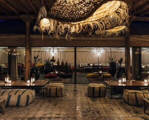 A cozy interior with a high ceiling, rustic wooden beams, and a central hanging light fixture. Tables and benches are arranged for dining or socializing. - Caravan by Habitas Agafay (Agafay)