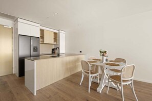 A modern kitchen with white cabinetry, a built-in island, and a dining area with wicker chairs and a white table. - Sea Glimpse Escape at Sandbar Burleigh (Gold Coast)