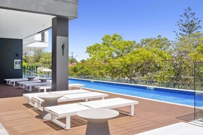 A poolside area with white lounge chairs and tables, a clear blue pool, and a view of trees and buildings in the background. - Sea Glimpse Escape at Sandbar Burleigh (Gold Coast)