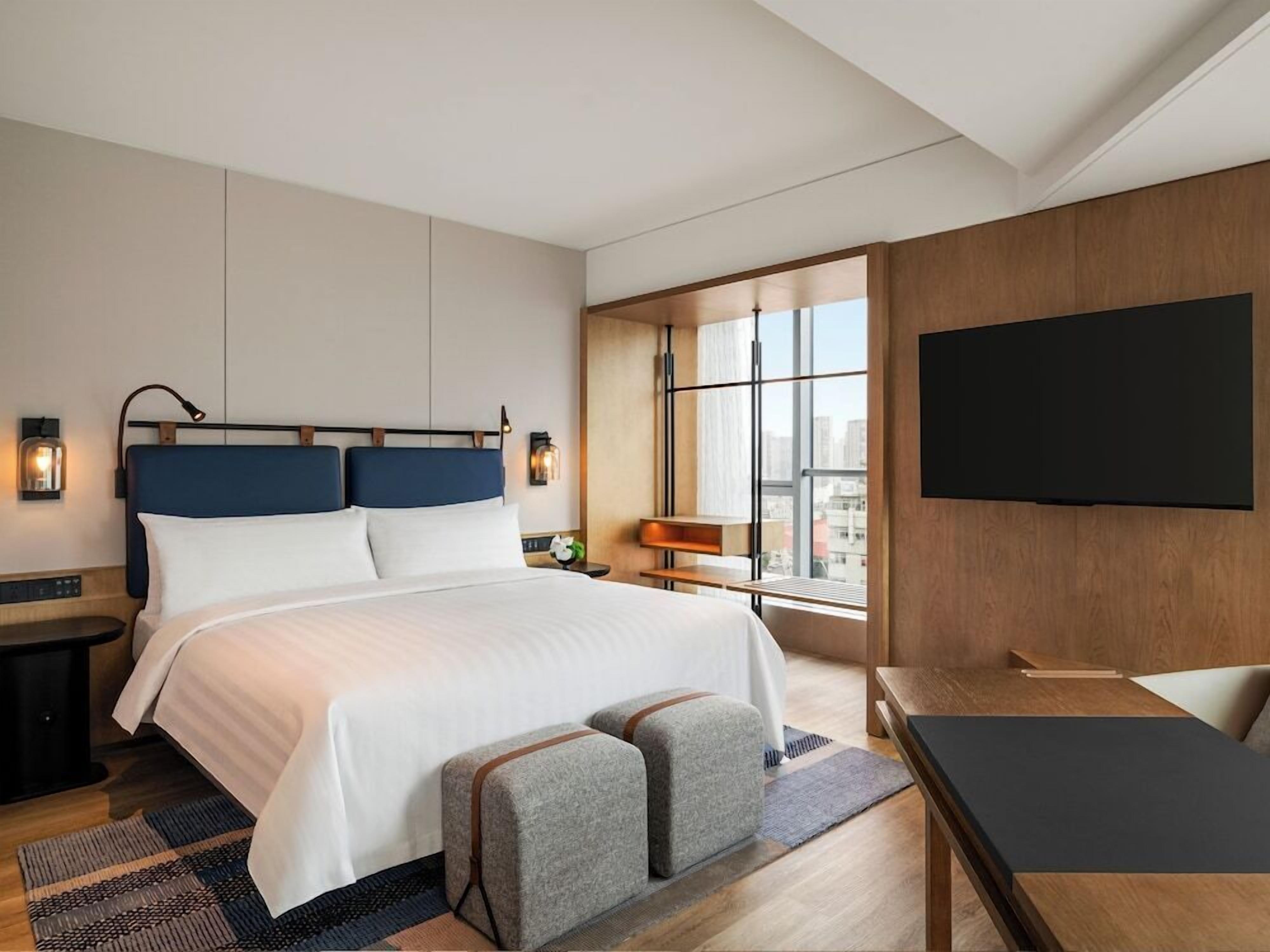 A hotel room with a bed, bedside tables, a TV, and a window with a city view.