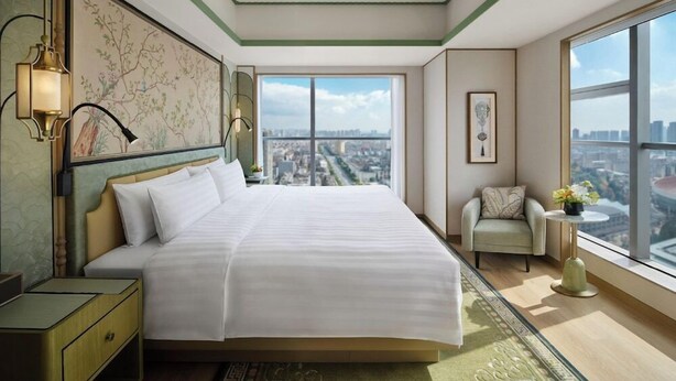 A hotel room with a large bed, a view of the city, a chair, and a small table with flowers. - JEN Kunming by Shangri-la (Kunming)