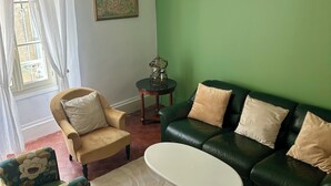 Ein Wohnzimmer mit grüner Wand, einer schwarzen Deckenleuchte, einem grünen Ledersofa mit beige farbenen Kissen, einem gelben Sessel und einem weißen Couchtisch, sowie einem gemusterten Sessel.