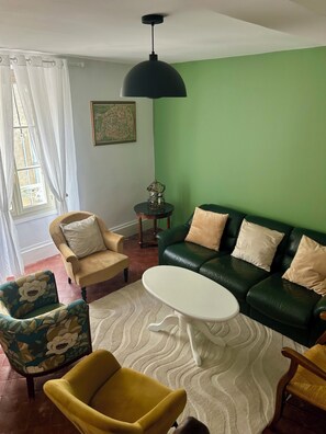 Ett vardagsrum med en grön vägg, en svart taklampa, en grön lädersoffa med beige kuddar, en gul fåtölj, ett vitt soffbord och en mönstrad fåtölj.