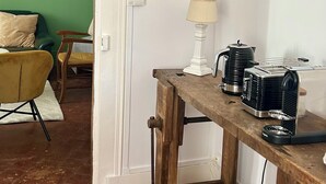 Ein Zimmer mit einem Holztisch, der eine Lampe, einen Wasserkocher und einen Toaster beherbergt. Eine grüne Wand und eine Tür zu einem anderen Raum sind ebenfalls zu sehen.