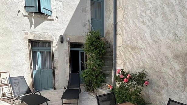 Ein Außenbereich mit einer Terrasse, einem Tisch und Stühlen, einer überdachten Sitzgruppe und einem Gebäude mit Steinverkleidung.