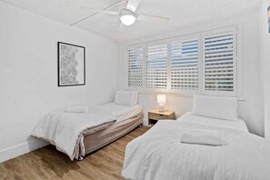 Ein Schlafzimmer mit zwei Betten, einem Deckenventilator, einem Nachttisch mit Lampe und einem Fenster mit Jalousien.