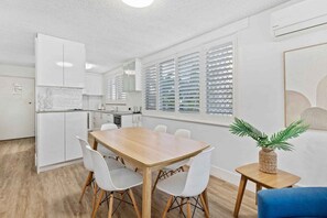 A modern kitchen with white cabinetry, a wooden dining table with white chairs, and a small side table with a plant. - Wyuna Coastal Escape - 3BR Apartment (Gold Coast)