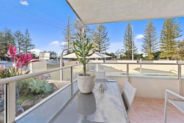 Ein Balkon mit Glastisch, einem Blumentopf und Blick auf ein Schwimmbecken und Bäume.
