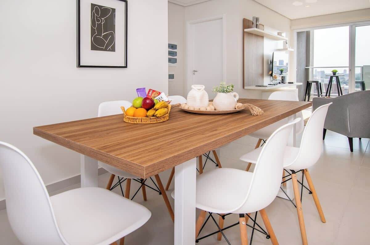 Un comedor moderno con una mesa de madera, sillas blancas, una cesta de fruta y una jarra sobre la mesa.