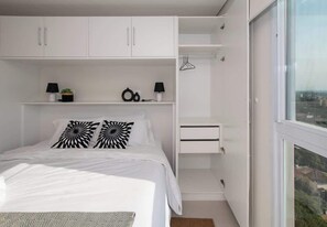 Un dormitorio con cama, armarios blancos, un estante pequeño con dos lámparas y vistas por una ventana.