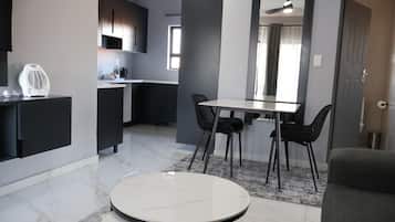 A modern living room with a round table, two black chairs, a grey sofa, a kitchen area with a dining table, and a ceiling fan.