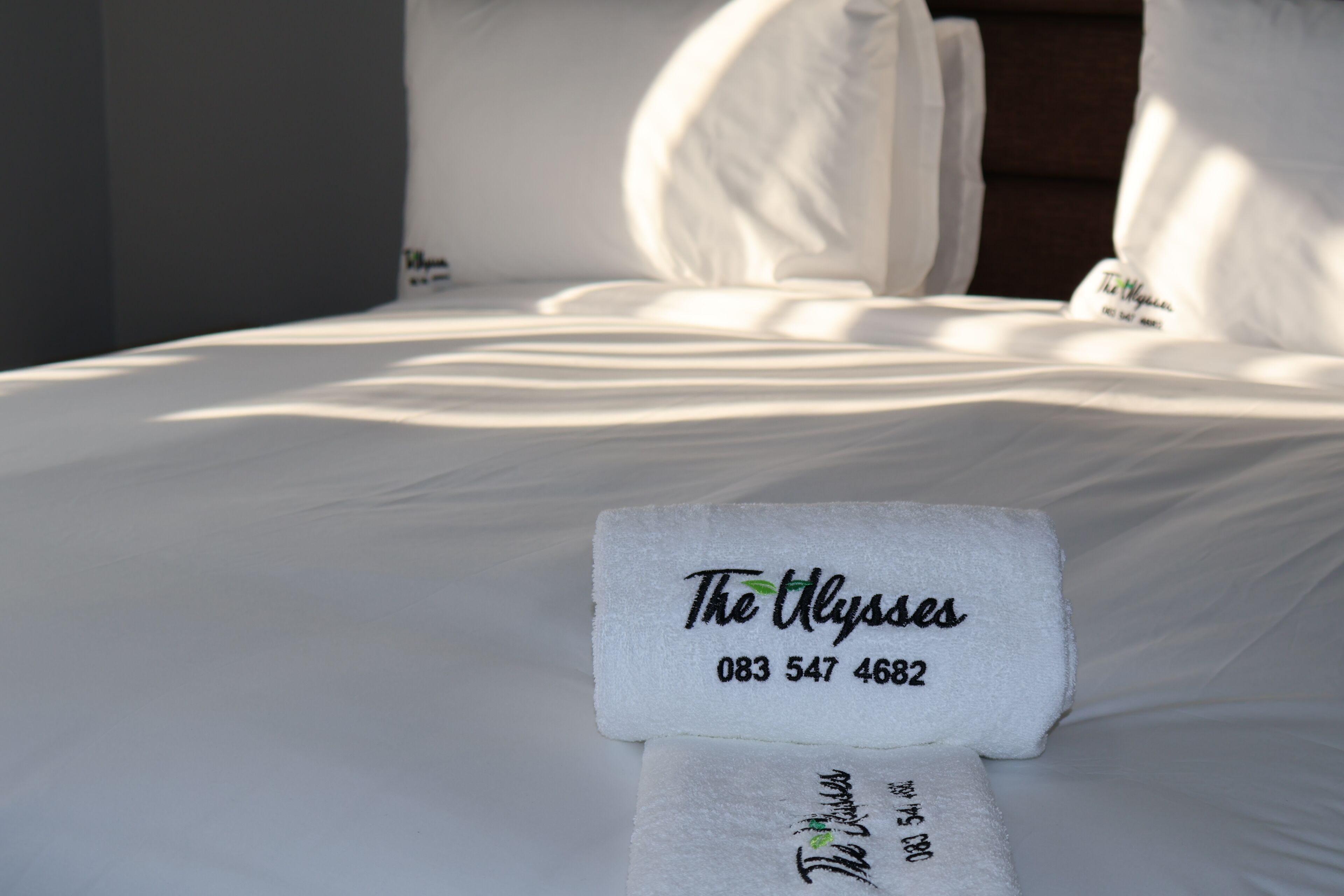 Ein ordentlich bezogenes Bett mit weißen Laken und einem Handtuch, das den Hotelnamen 'The Alyses' und die Telefonnummer zeigt.