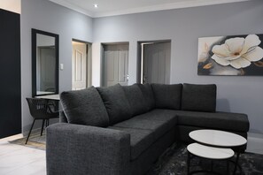 A modern living room with a grey sectional sofa, two round tables, and a large floral painting on the wall. - The Ulysses Motel (Polokwane)