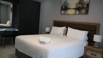 A hotel room with a large bed, a desk, a chair, a nightstand with a lamp, and a painting on the wall.