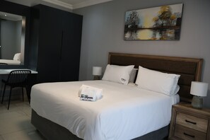 Quarto de hotel com uma cama grande, uma mesa de escritório, uma cadeira, uma mesa de cabeceira com uma luminária e um quadro na parede.