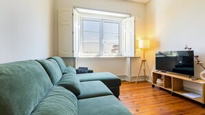 A living room with a green sofa, a TV on a wooden stand, a lamp, and a window with curtains. - GuestReady-Charming 3-Bedroom Lisbon Retreat (Lisbon)