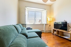 A living room with a green sofa, a TV on a wooden stand, a lamp, and a window with curtains. - GuestReady-Charming 3-Bedroom Lisbon Retreat (Lisbon)
