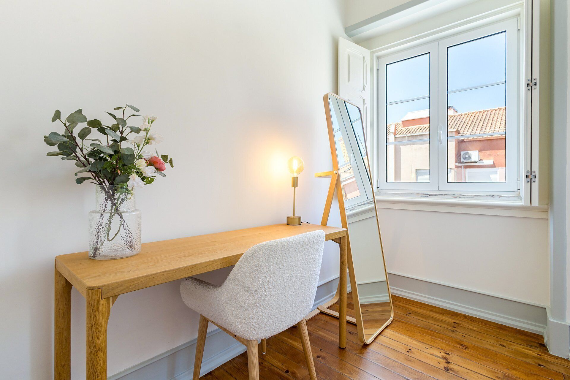 Una habitación con un escritorio de madera, una silla blanca, un florero con flores, una lámpara de pie y una ventana que ofrece vistas a edificios.