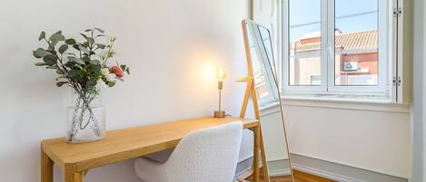 A room with a wooden desk, a white chair, a vase with flowers, a standing lamp, and a window with a view of buildings.