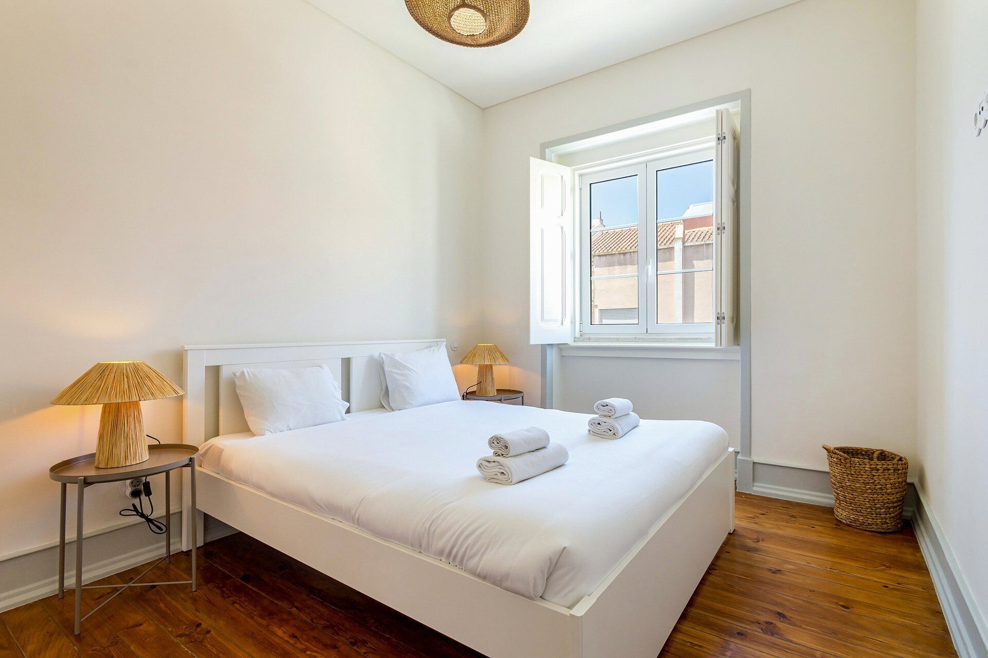 Una cama bien hecha con sábanas blancas y dos toallas enrolladas, una mesita de noche con una lámpara, una ventana con vistas a edificios y una cesta tejida en el suelo.