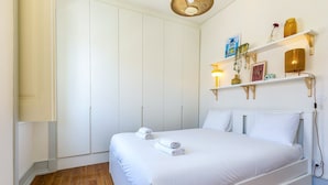 A bedroom with a bed, white walls, wooden flooring, a wardrobe, and a shelf with decorative items. - GuestReady-Charming 3-Bedroom Lisbon Retreat (Lisbon)