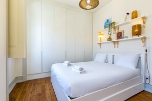 A bedroom with a bed, white walls, wooden flooring, a wardrobe, and a shelf with decorative items. - GuestReady-Charming 3-Bedroom Lisbon Retreat (Lisbon)