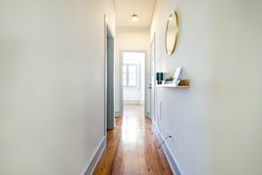 A hallway with a wooden floor, white walls, a door at the end, and a wall-mounted shelf with a lamp and a small plant. - GuestReady-Charming 3-Bedroom Lisbon Retreat (Lisbon)