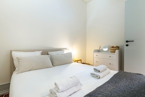 A neatly made bed with white linens, a bedside table with a lamp, a small dresser, and a door to the right. - GuestReady-Charming 3-Bedroom Lisbon Retreat (Lisbon)