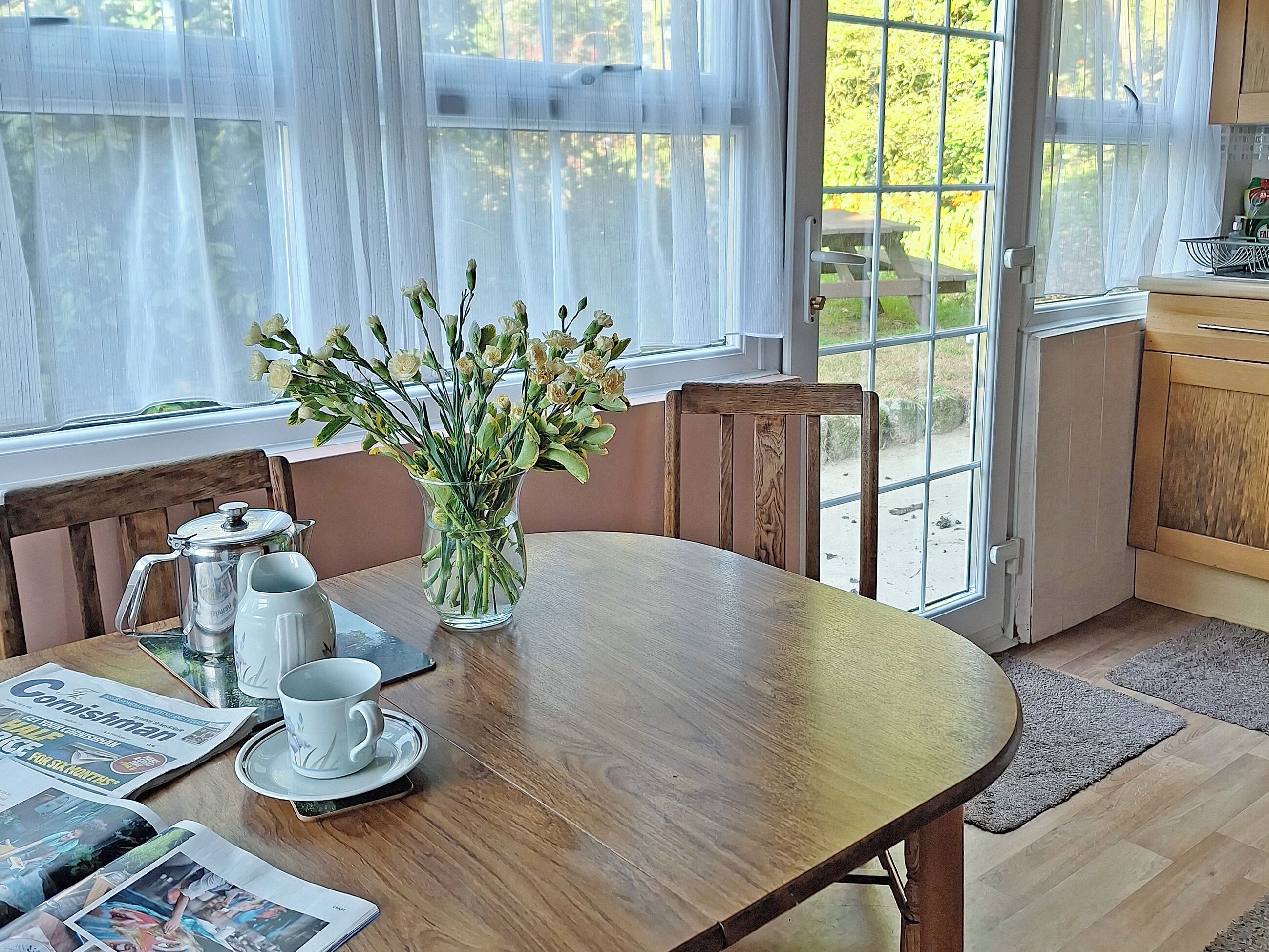Une table de salle à manger en bois, ornée d’un vase de fleurs, d’une cafetière et d’un journal.