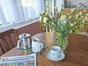 A dining table with a newspaper, a coffee pot, a cup, saucer, and a vase with flowers. - The Cottage @ Tredinney Farm - UK52162 (St Buryan)