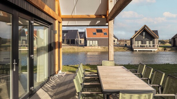 A covered outdoor area with a table and chairs, a view of a water body, and neighboring houses. - Haven Near Nieuwvliet-bad (Nieuwvliet)