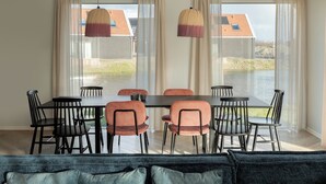 A dining area with a table and chairs, large windows with curtains, and a view of a building with a red roof. - Haven Near Nieuwvliet-bad (Nieuwvliet)