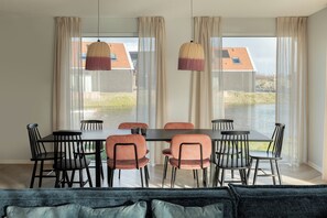 A dining area with a table and chairs, large windows with curtains, and a view of a building with a red roof. - Haven Near Nieuwvliet-bad (Nieuwvliet)