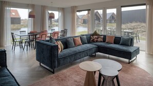 A modern living room with a large sectional sofa, a dining area with chairs, and large windows offering views of a water body and buildings. - Haven Near Nieuwvliet-bad (Nieuwvliet)