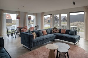 A modern living room with a large sectional sofa, a dining area with chairs, and large windows offering views of a water body and buildings. - Haven Near Nieuwvliet-bad (Nieuwvliet)