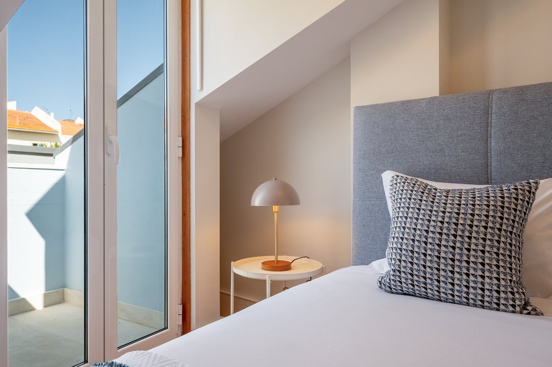 Une chambre d’hôtel avec un lit, une table de chevet avec une lampe, un coussin à motifs et une vue à travers une porte vitrée.