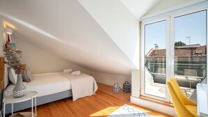 Ett modernt sovrum i loftstil med ett sluttande tak, ett stort fönster, en säng, en gul stol och ett litet bord med en vas.