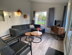 A modern living room with a grey sofa, a wooden coffee table, a TV stand, and a view of the outdoors. - Spacious Bungalow Near Oostkapelle for 4 (Oostkapelle)