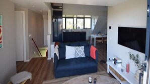 A modern living room with a dark blue sofa, a flat-screen TV, and a kitchen area visible through an open door. - Coastal Apartment in Oostkapelle (Oostkapelle)